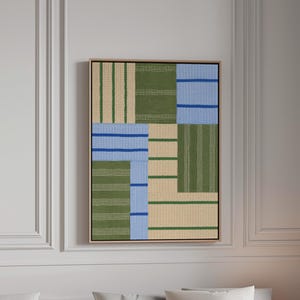 May include: A rectangular wall art piece featuring a geometric patchwork design of textured fabric panels in olive green, light blue, and beige. The composition includes alternating vertical and horizontal stripes, framed in a thin light wood border.