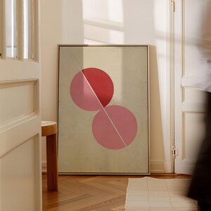 May include: Abstract art print featuring two overlapping pink circles, one red and one lighter pink, bisected by diagonal white lines. The artwork is framed in a thin gold frame and leans against a wall in a room with a wooden floor.