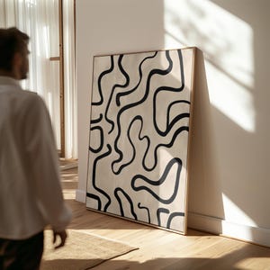 Può includere: Grande stampa artistica astratta incorniciata. L'opera presenta linee nere e fluide su uno sfondo color crema. La cornice è di colore legno chiaro. Il pezzo è appoggiato a una parete bianca.
