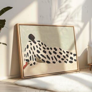 May include: A framed wall art print featuring a stylised figure reclining against a pale green and beige background. The figure wears a white garment covered in large, irregular black brushstroke spots, with one visible hand painted in a solid red tone.