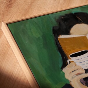 May include: Abstract painting in a natural wood frame. The artwork features a person holding a white coffee cup with blue stripes. The background is green with black and tan accents. The painting is on a wooden surface.