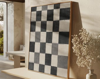 May include: A framed artwork featuring a checkerboard pattern in shades of black, gray, and off-white. The artwork is displayed on a light-colored wooden shelf, with a vase of white flowers and a small wooden dish nearby. The frame is a light brown color.