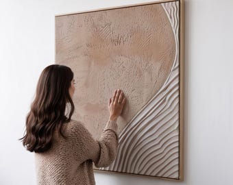 Op de afbeelding: Een groot, vierkant, getextureerd wandkunstwerk met een lichtbruin en wit ontwerp. Het kunstwerk heeft een cirkelvorm en gelaagde, golfachtige patronen aan de rechterkant. Het stuk is omlijst in licht hout.