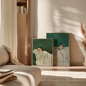 May include: Two framed art prints featuring illustrations of people. One shows a person resting, wrapped in a striped blanket. The other depicts a person holding a cup. Both have a green background and gold frames.