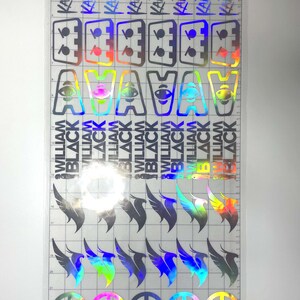 May include: A sheet of holographic stickers with various designs. The stickers include text that reads "KAI WACHI" and "WILLIAM BLACK", along with symbols such as eyes, wings, and peace signs. The stickers are on a clear backing.