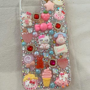 Puede incluir: Funda de tel&eacute;fono transparente decorada con pedrer&iacute;a rosa, roja y transparente, y varios abalorios. Los abalorios incluyen figuras de Hello Kitty, un lazo, una estrella, una fresa, una piruleta, una magdalena y un helado.