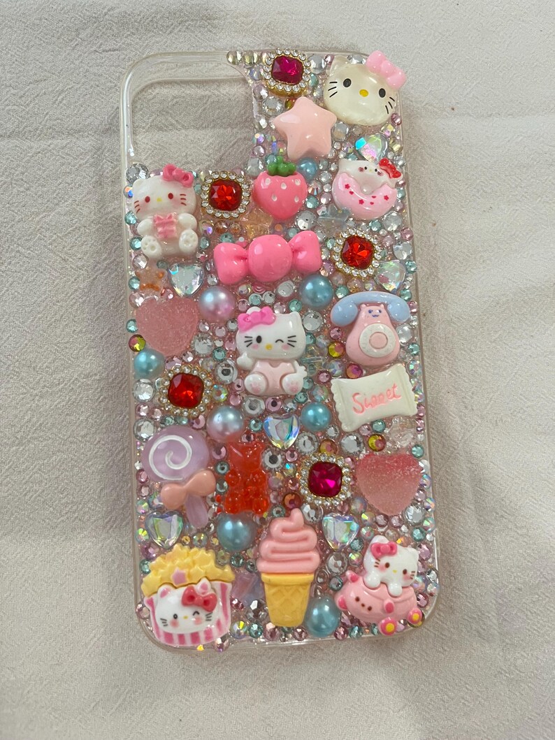 Puede incluir: Funda de tel&eacute;fono transparente decorada con pedrer&iacute;a rosa, roja y transparente, y varios abalorios. Los abalorios incluyen figuras de Hello Kitty, un lazo, una estrella, una fresa, una piruleta, una magdalena y un helado.