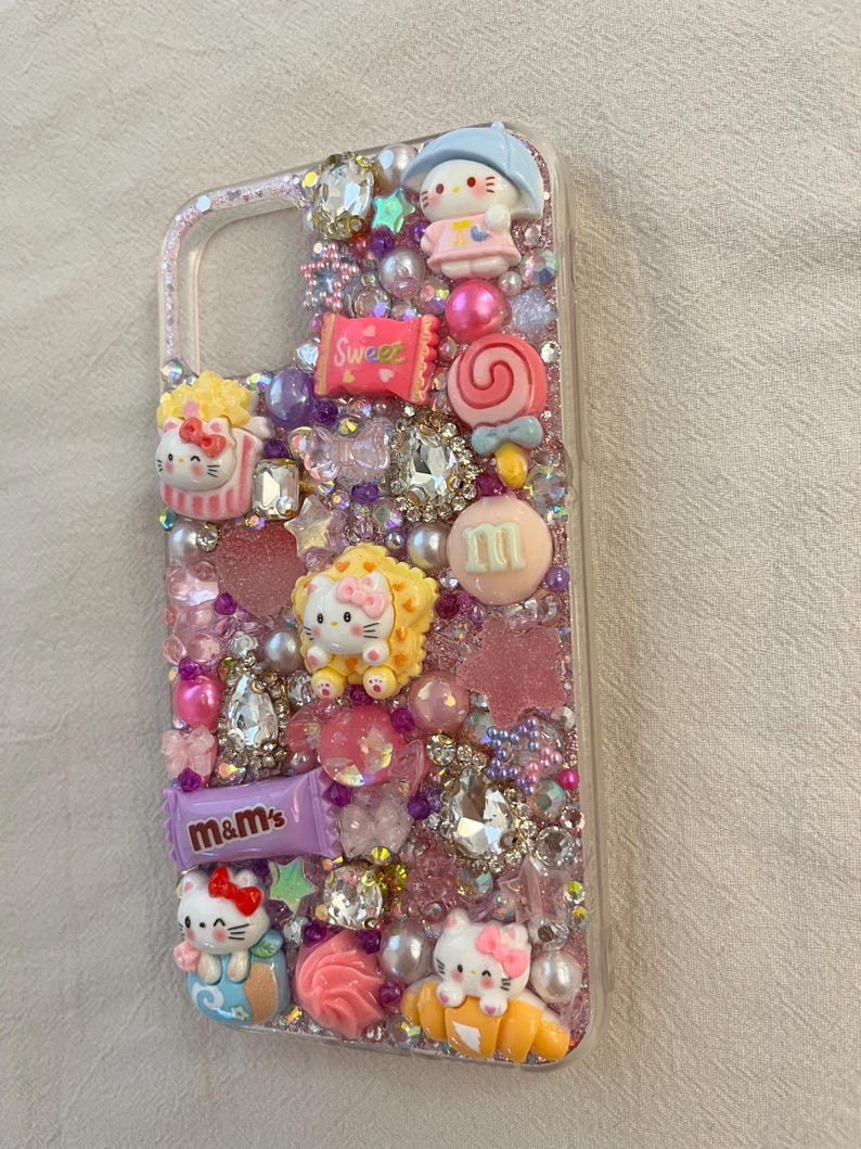Pu&ograve; includere: Una custodia per telefono rosa decorata con strass, glitter e vari ciondoli, tra cui personaggi di Hello Kitty, caramelle e M&M's.