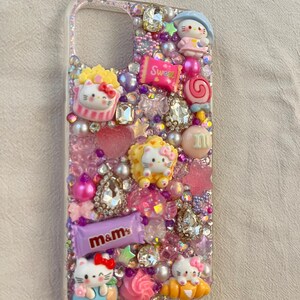 Pu&ograve; includere: Una custodia per telefono trasparente decorata con strass, glitter e vari ciondoli, tra cui personaggi di Hello Kitty, caramelle e cuori. La custodia &egrave; progettata in uno schema di colori rosa e viola.