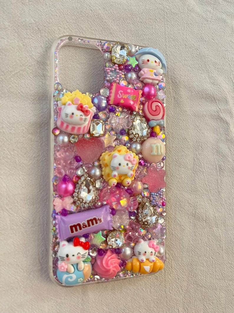 Pu&ograve; includere: Una custodia per telefono trasparente decorata con strass, glitter e vari ciondoli, tra cui personaggi di Hello Kitty, caramelle e cuori. La custodia &egrave; progettata in uno schema di colori rosa e viola.