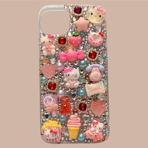 Puede incluir: Una funda transparente para tel&eacute;fono adornada con brillantes pedrer&iacute;a y varios dijes rosas, rojos y amarillos. Los dijes incluyen figuras de Hello Kitty, corazones, lazos y un tel&eacute;fono. La palabra "Sweet" est&aacute; escrita en una peque&ntilde;a pancarta.