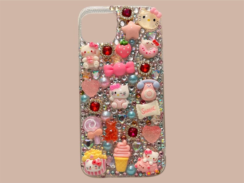 Puede incluir: Una funda transparente para tel&eacute;fono adornada con brillantes pedrer&iacute;a y varios dijes rosas, rojos y amarillos. Los dijes incluyen figuras de Hello Kitty, corazones, lazos y un tel&eacute;fono. La palabra "Sweet" est&aacute; escrita en una peque&ntilde;a pancarta.