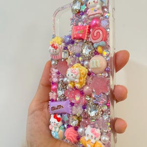 Pu&ograve; includere: Una custodia per telefono trasparente decorata con glitter rosa, viola e bianco, strass e ciondoli. I ciondoli includono personaggi di Hello Kitty, caramelle e una barretta di cioccolato "m&m's".
