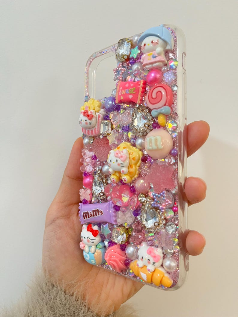 Pu&ograve; includere: Una custodia per telefono trasparente decorata con glitter rosa, viola e bianco, strass e ciondoli. I ciondoli includono personaggi di Hello Kitty, caramelle e una barretta di cioccolato "m&m's".