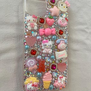 Puede incluir: Funda de tel&eacute;fono transparente decorada con varios abalorios rosas y rojos, incluyendo figuras de Hello Kitty, corazones, lazos y pedrer&iacute;a. Otros abalorios incluyen una fresa, un helado y un tel&eacute;fono. La palabra "Sweet" est&aacute; escrita en un peque&ntilde;o rect&aacute;ngulo blanco.