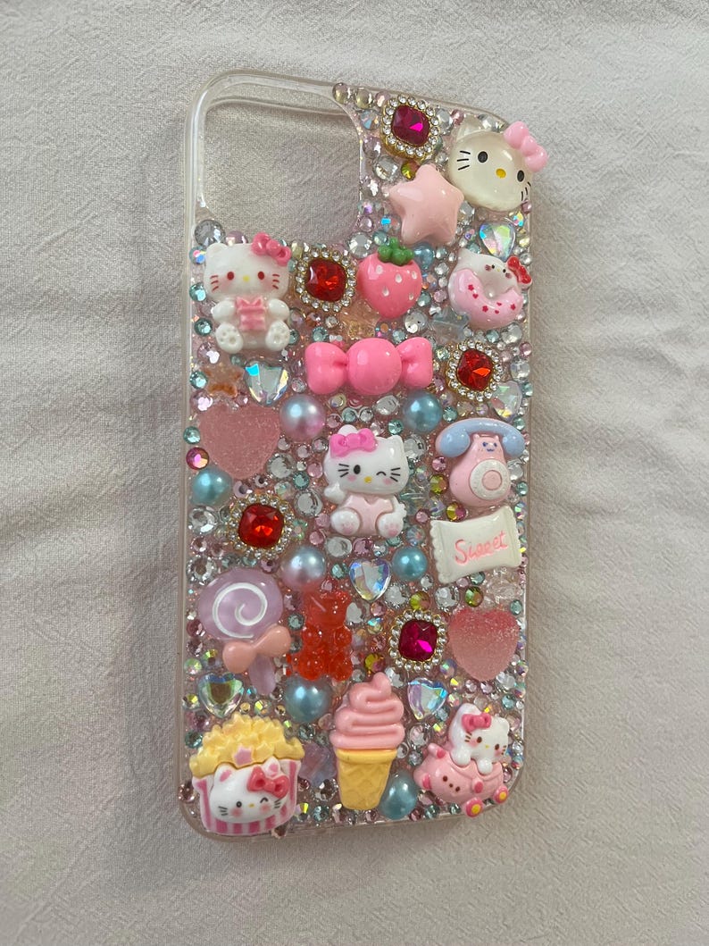 Puede incluir: Funda de tel&eacute;fono transparente decorada con varios abalorios rosas y rojos, incluyendo figuras de Hello Kitty, corazones, lazos y pedrer&iacute;a. Otros abalorios incluyen una fresa, un helado y un tel&eacute;fono. La palabra "Sweet" est&aacute; escrita en un peque&ntilde;o rect&aacute;ngulo blanco.