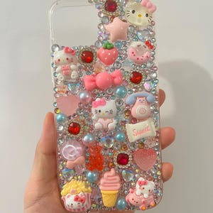 Puede incluir: Funda de tel&eacute;fono transparente decorada con varios adornos rosas, rojos y transparentes. Presenta figuras de Hello Kitty, una fresa, un lazo, una magdalena, un helado y la palabra "Sweet". La funda est&aacute; adornada con pedrer&iacute;a y perlas.