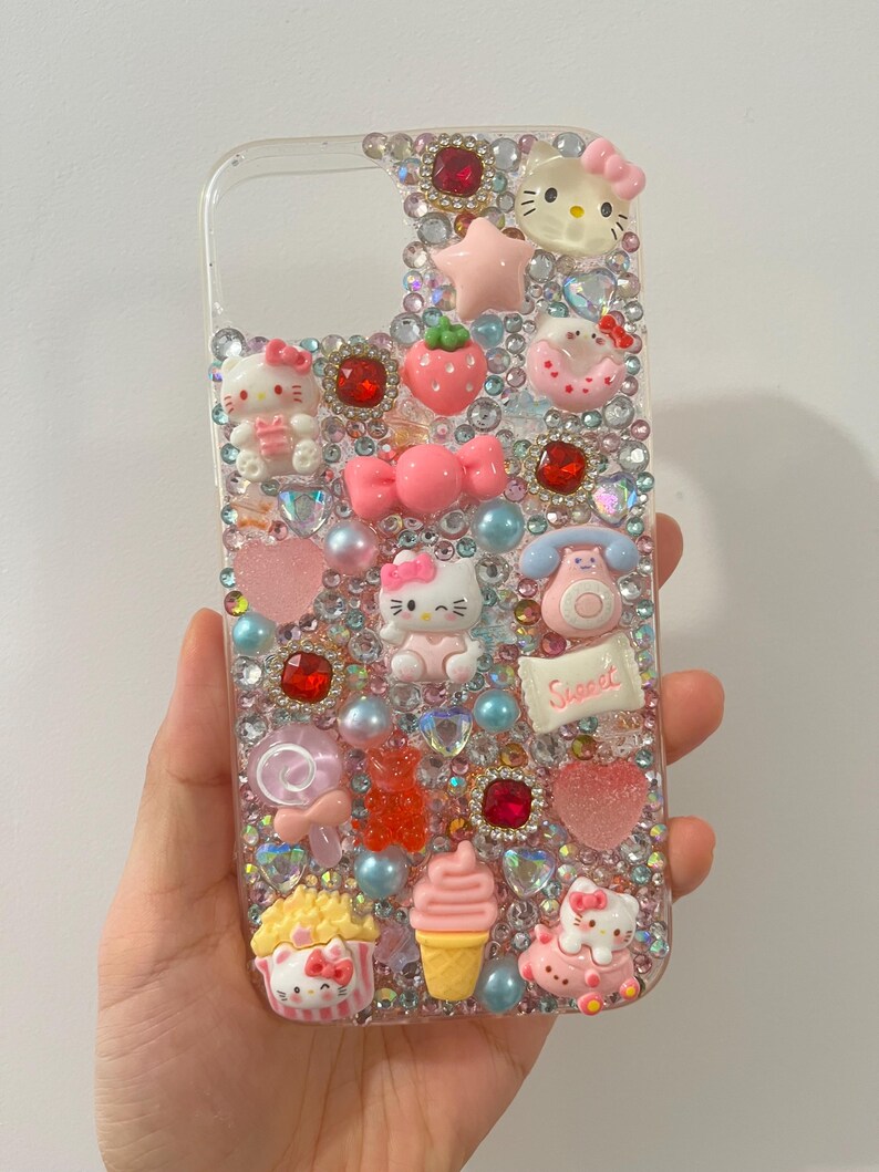 Puede incluir: Funda de tel&eacute;fono transparente decorada con varios adornos rosas, rojos y transparentes. Presenta figuras de Hello Kitty, una fresa, un lazo, una magdalena, un helado y la palabra "Sweet". La funda est&aacute; adornada con pedrer&iacute;a y perlas.