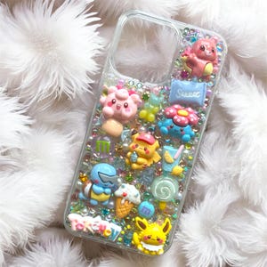 Könnte beinhalten: Transparente Handyhülle mit bunten Zeichentrickfiguren, Süßigkeiten und Perlen verziert. Die Hülle zeigt verschiedene Figuren, darunter ein gelbes Pikachu, ein rosa Clefairy und ein blaues Schiggy. Das Wort "Pokemon" ist unten sichtbar.