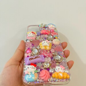 Pu&ograve; includere: Una custodia per telefono trasparente decorata con glitter rosa, viola e bianco, strass e ciondoli. I ciondoli includono personaggi di Hello Kitty, una canna di zucchero, un croissant e una barretta di cioccolato M&M's.