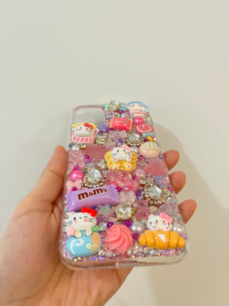 Pu&ograve; includere: Una custodia per telefono trasparente decorata con glitter rosa, viola e bianco, strass e ciondoli. I ciondoli includono personaggi di Hello Kitty, una canna di zucchero, un croissant e una barretta di cioccolato M&M's.