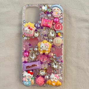 Pu&ograve; includere: Una custodia per telefono trasparente con uno sfondo di glitter rosa, viola e bianco. La custodia &egrave; decorata con personaggi di Hello Kitty, caramelle e strass. Il testo "Sweet" e "m&m's" &egrave; visibile sulla custodia.