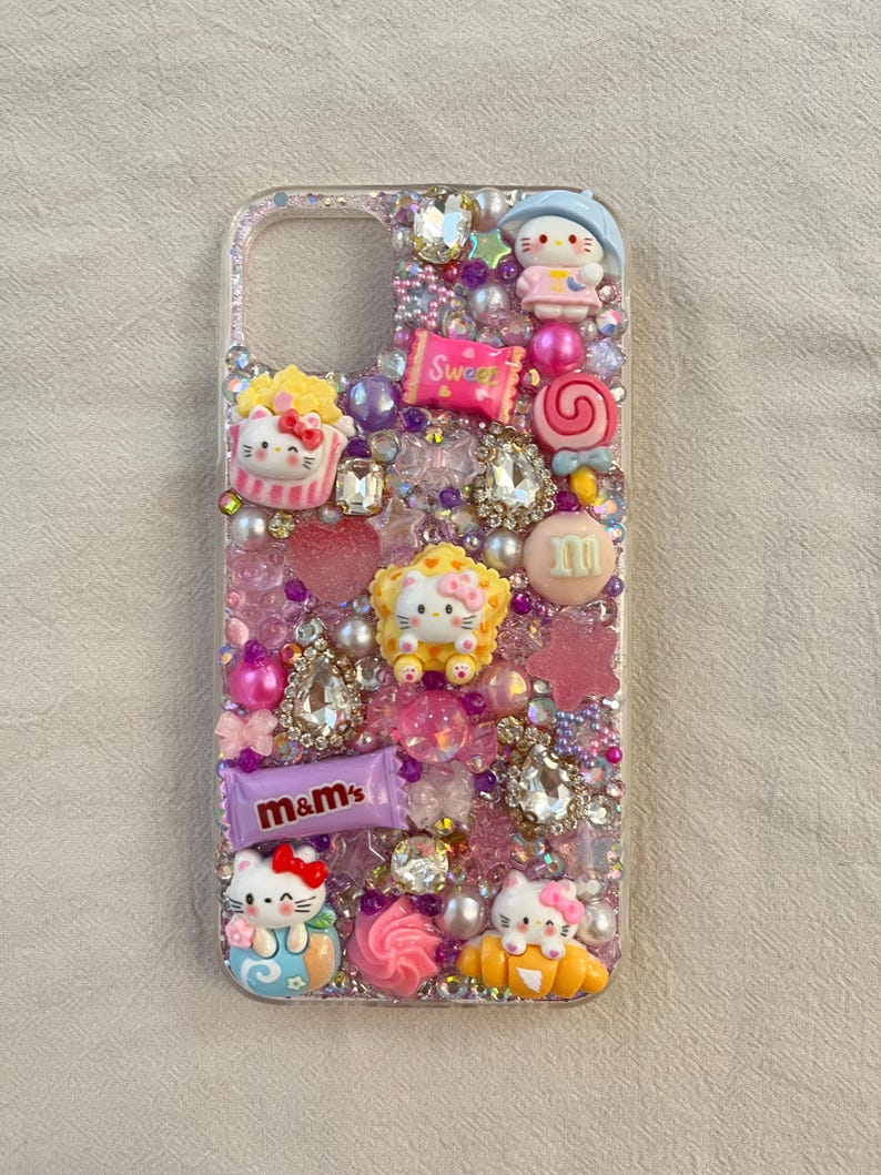 Pu&ograve; includere: Una custodia per telefono trasparente con uno sfondo di glitter rosa, viola e bianco. La custodia &egrave; decorata con personaggi di Hello Kitty, caramelle e strass. Il testo "Sweet" e "m&m's" &egrave; visibile sulla custodia.