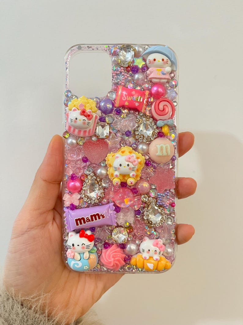 Pu&ograve; includere: Una custodia per telefono trasparente decorata con glitter rosa, viola e bianco, strass e vari ciondoli, tra cui personaggi Hello Kitty, caramelle e una barretta di cioccolato M&M's.