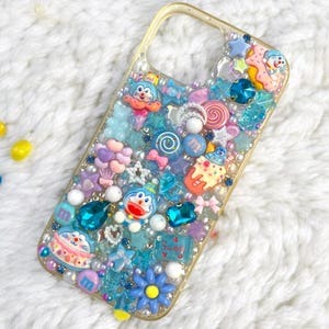 Può includere: Una custodia per telefono trasparente decorata con ciondoli colorati, tra cui caramelle blu e rosa, stelle e personaggi dei cartoni animati. La custodia è ricoperta di glitter e strass.