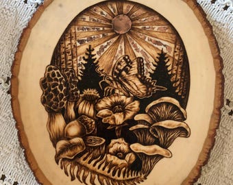 May include: A wood slice with a woodburned design of a butterfly, mushrooms, flowers, and a sun with rays. The design is in shades of brown and black.