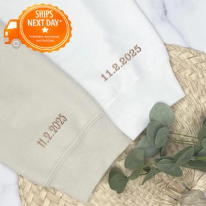 May include: Two sweatshirts, one cream and one white, are displayed. Each sleeve cuff features embroidered text reading "11.2.2025" in a brown thread. The image includes a "SHIPS NEXT DAY" graphic with an orange truck.