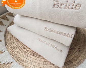 Puede incluir: Tres sudaderas beige dobladas con texto bordado: "Bride", "Bridesmaid" y "Maid of Honor". Las sudaderas están apiladas sobre una estera de paja tejida. La imagen incluye un gráfico de "Envío al día siguiente".