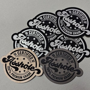 Puede incluir: Un conjunto de parches redondos con la palabra "Asshole" en una fuente estilizada. Cada parche presenta el texto "CERTIFIED" y "100% PREMIUM GRADE" en un diseño circular. Los parches son de color negro, plateado, beige y gris.
