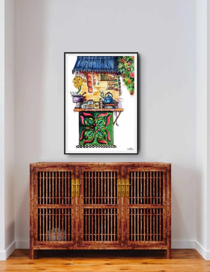 May include: Watercolor painting of a street food stall with a blue awning and green and red accents. The stall features a table with various items, including a teapot and jars. The artwork is framed and displayed above a wooden cabinet.