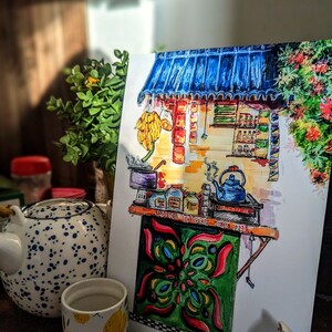 May include: Watercolor illustration of a tea stall with a blue awning, bananas, and various jars. The stall has a green and red patterned front. A blue teapot and a cup with lemons are in the foreground.