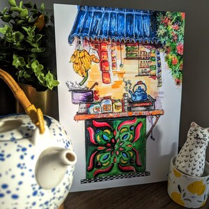 May include: A watercolor painting of a street food stall with a blue awning, bananas, and various items for sale. The stall has a green and red decorative panel. A blue and white teapot and a cat figurine are in the foreground.