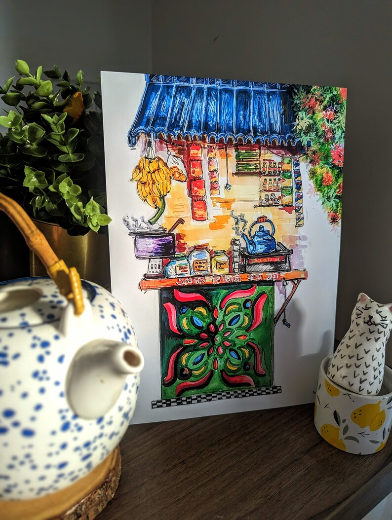 May include: A watercolor painting of a street food stall with a blue awning, bananas, and various items for sale. The stall has a green and red decorative panel. A blue and white teapot and a cat figurine are in the foreground.