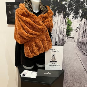 Op de afbeelding: Een oranje imitatiebont sjaal gedrapeerd over een zwarte top op een mannequin. De sjaal heeft een getextureerd uiterlijk en wordt gecombineerd met een geruite capuchon. Een bord met de merknaam en een afbeelding van de sjaal.