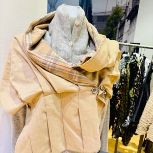 Op de afbeelding: Een beige, geplooid, kort jack met een geruite sjaal. Het jack heeft een knoopsluiting en wordt getoond op een mannequin.