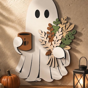 May include: A white ghost wall decoration featuring two black oval eyes, holding a brown mug with a pumpkin icon in one hand and a bouquet of dried-style foliage in shades of green, tan, and brown in the other hand, with layered cut-out details.