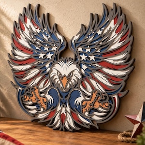 Puede incluir: Una pieza de arte mural de madera multicapa que representa un águila calva con las alas extendidas. El diseño incorpora colores rojo, blanco y azul con patrones de estrellas en las alas, un pico naranja y garras amarillas en relieve.