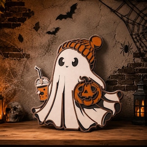 May include: A layered wooden cutout of a white ghost wearing an orange beanie, holding a brown iced coffee drink in one hand and a carved orange jack-o'-lantern pumpkin in the other, featuring dark outlines and a multi-dimensional design.