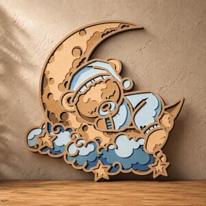Könnte beinhalten: Ein mehrschichtiges Wandbild aus Holz, das einen schlafenden Bären mit Schlafmütze auf einer Mondsichel zeigt. Das Design umfasst hell- und dunkelblaue Wolken sowie zwei kleine Sterne, die alle mit ausgeprägter Tiefe und Textur gestaltet sind.