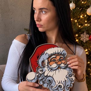 May include: A layered wooden wall art piece depicting the face of Santa Claus with a red hat, a white mustache, and a detailed beard. The design features stacked cutouts in shades of red, white, light blue, and dark gray, creating a dimensional effect.