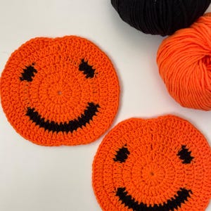May include: Two orange crocheted pumpkin coasters with black yarn smiley faces. The coasters are on a white surface with orange and black yarn balls.