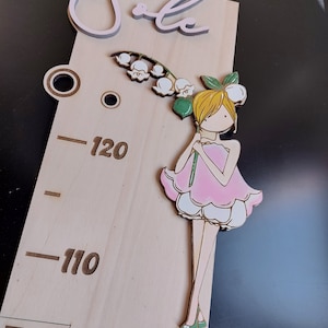 Può includere: Grafico di altezza in legno con un design a fata. Il grafico presenta una fata rosa e bianca che tiene un fiore, con misure in centimetri. La parola "Sale" è in alto.
