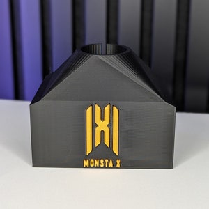 May include: Black 3D-printed stand with a gold logo and the text "MONSTA X". The stand has a geometric design with a cylindrical top. The logo is a stylized emblem.