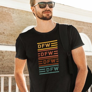 Peut inclure: Un t-shirt noir avec un design graphique rétro représentant le texte "DFW" en format empilé avec des rayures colorées.