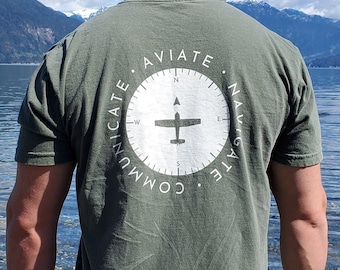 Puede incluir: Una camiseta de manga corta color verde oliva con un gráfico circular blanco en la espalda. El diseño incluye una rosa de los vientos, la silueta de un avión y el texto "AVIATE NAVIGATE COMMUNICATE" impreso en disposición circular.