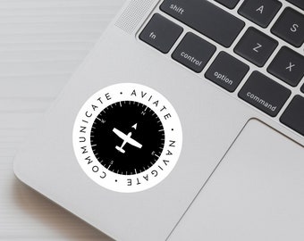 Könnte beinhalten: Ein schwarz-weißer Aufkleber mit einem Kompass und einem Flugzeugdesign. Der Aufkleber lautet "Kommunizieren, Fliegen, Navigieren".